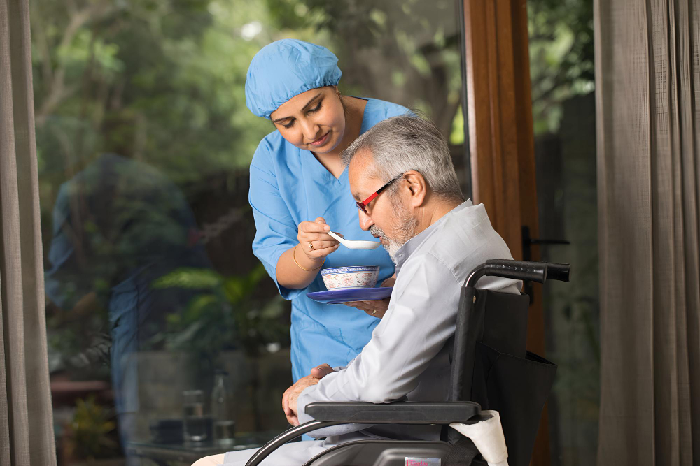 A female caregiver in a blue uniform feeding an elderly man sitting in a wheelchair